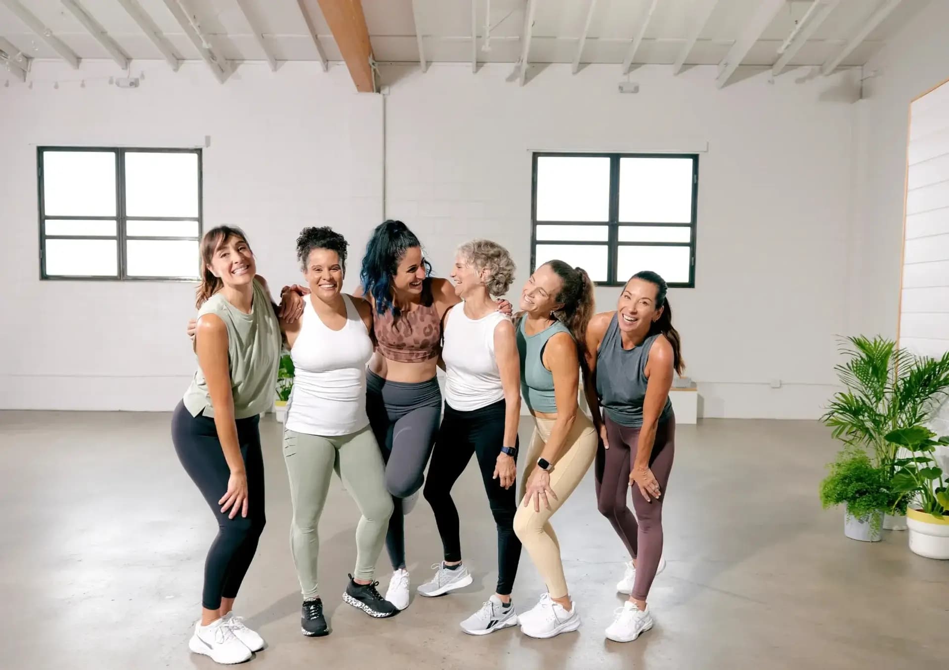 Six women in activewear are standing together in a light-filled studio, smiling and laughing. They appear to be in a post-workout moment, with casual poses and genuine expressions of joy and camaraderie. White walls and large windows create a bright, inviting atmosphere.