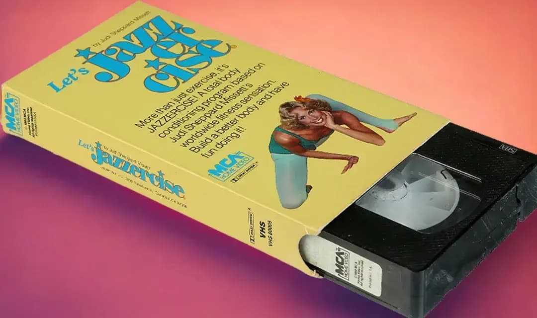 A vintage VHS tape of "Let’s Jazzercise" is shown with a bright yellow cover. It features an illustrated woman in workout attire, posing energetically. The background is a gradient of pink and orange hues, highlighting the retro exercise theme.