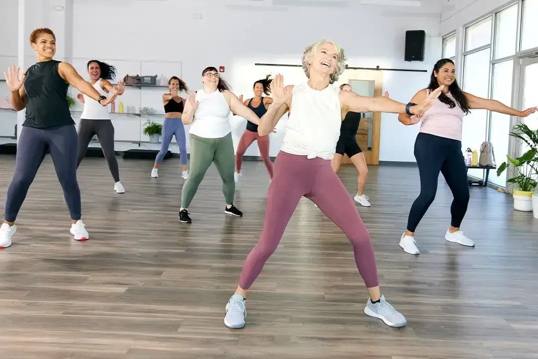 A diverse group of women smile and exercise together in a bright, spacious fitness studio, all wearing athletic clothing and moving energetically with arms outstretched.