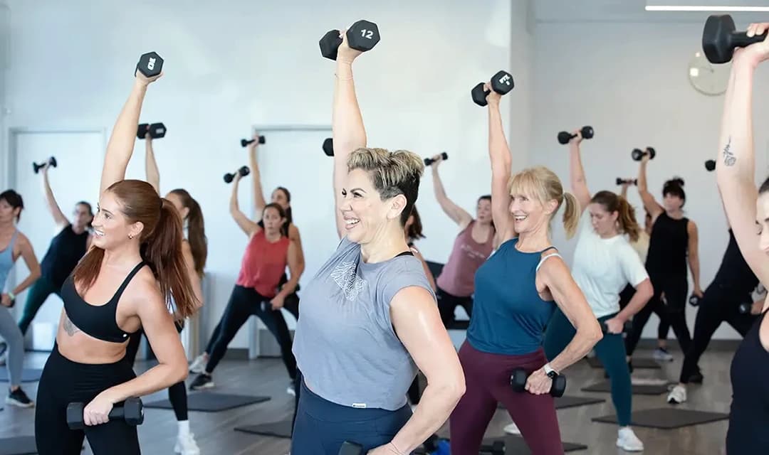 A group exercise class with diverse individuals lifting dumbbells. They are smiling and raising one arm with a dumbbell overhead. The setting appears bright and spacious, with various gym equipment visible in the background.
